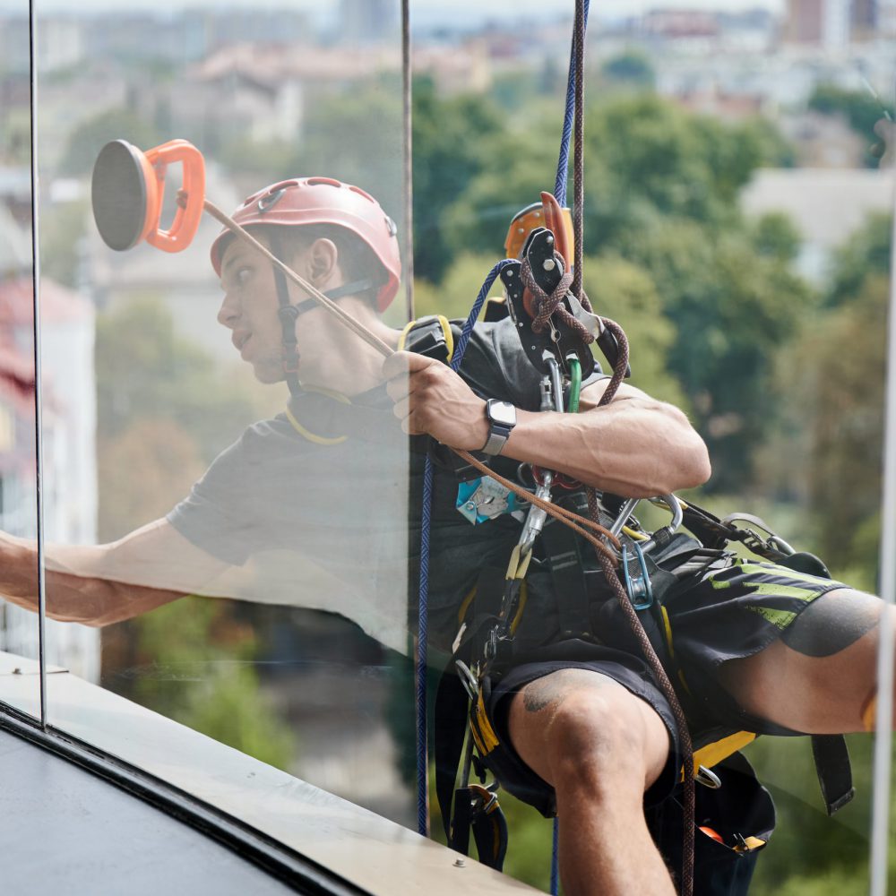 Industrial mountaineering cleaning service worker hanging on rope and wiping window. View from inside building. Cleaner using safety lifting equipment while cleaning glass of high-rise building.